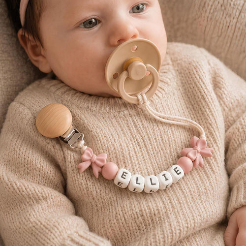 Baby wearing a personalized bow pacifier clip with a name in white letter beads and pink silicone bows