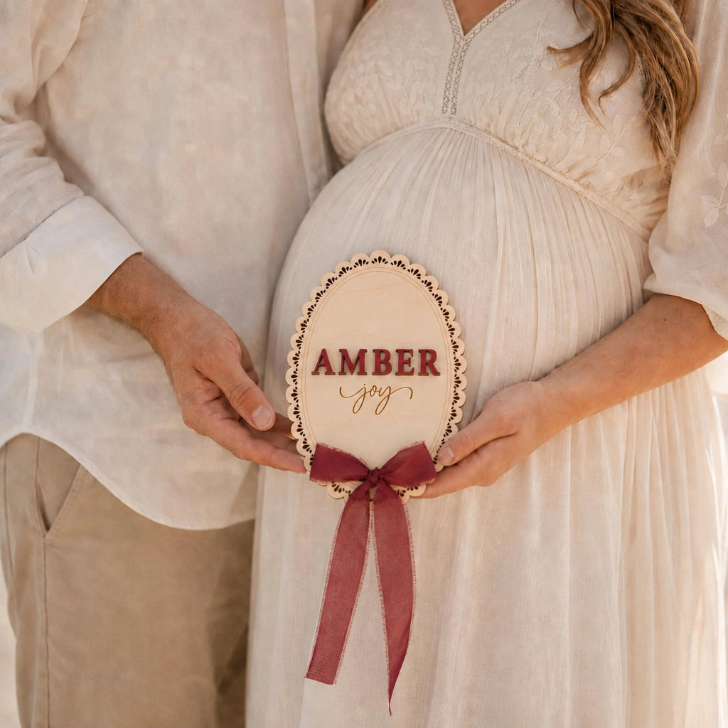 Baby name photo prop held at a maternity session, scalloped plaque with a wine ribbon