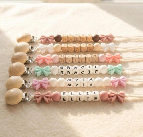 Four personalized bow pacifier clips in a flat lay, each with a baby name in letter beads and a matching bow